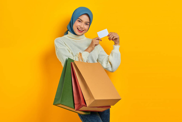 Весела жінка з Азії у білому светрі, що тримає сумочки і показує кредитну картку на жовтому фоні. - Фото, зображення