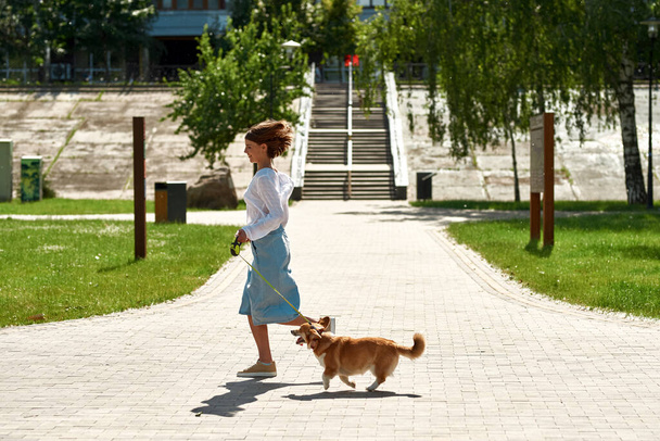Side view of pleased young caucasian girl running with adorable Corgi dog on sidewalk in park. Concept of relationship between human and animal. Idea of owner and pet friendship. Sunny summer day - Zdjęcie, obraz