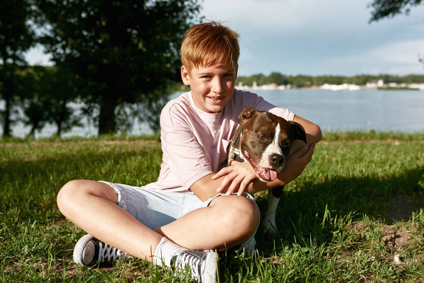 Smiling caucasian boy resting and hugging Staffordshire Bull Terrier dog on green meadow in park. Concept of relationship between human and animal. Idea of owner and pet friendship. Sunny summer day - Фото, зображення