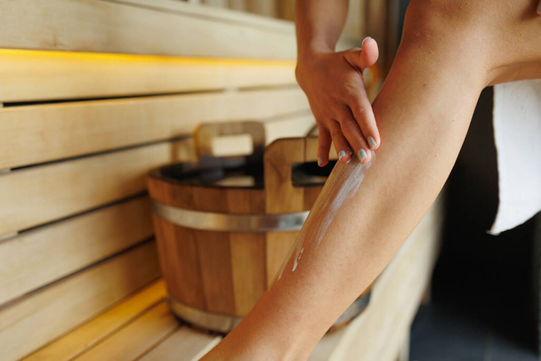 Vista de cerca de la mujer que aplica crema hidratante en la piel de las piernas en la sauna - Foto, Imagen