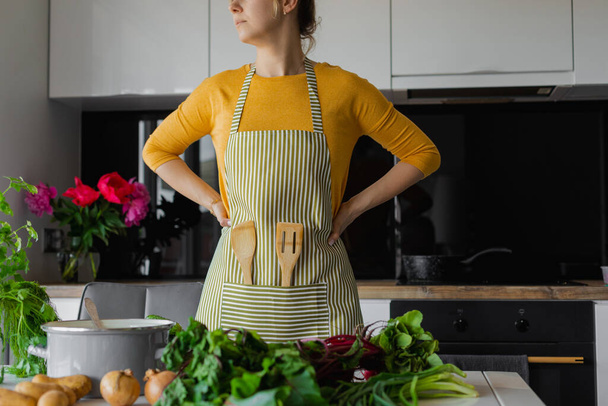 Cropped blonde woman in striped kitchen apron with wooden spatula utensil inside. Female arms akimbo, looking away, thinking what recipe to choose. Fresh vitamin vegetables as ingredients on the table - Photo, Image
