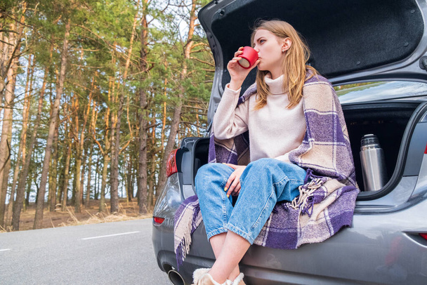 Nuori nainen istuu auton takakontissa kahvikupit ja termospullo puolella tietä metsässä lämpimänä syksyllä tai keväällä päivä. Sinun on aika mennä. Pohdinta ja lepo yksinäisyydessä - Valokuva, kuva