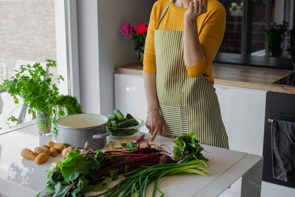 Nierozpoznawalna kobieta w fartuchu wybierająca warzywa do posiekania i krojenia na pokładzie w kuchni. Świeże organiczne ogórki rustykalne, ziemniaki, cebula, buraki jako składniki. Zdrowe jedzenie latem - Zdjęcie, obraz