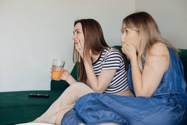 2人の陽気な女性がリビングルームの緑の快適なソファに座って、一緒にユーモラスなテレビ番組を見て、楽しい側の景色を楽しんでいます。女性の友情,家庭のリラクゼーションとレジャー,テレビ. - 写真・画像