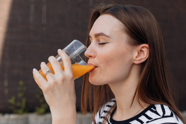 Портрет красивой молодой женщины, пьющей и глотающей свежий холодный апельсиновый сок на открытом воздухе на террасе с видом сбоку. Леди с закрытыми глазами наслаждается вкусом напитка, освежая в летний день. - Фото, изображение