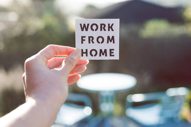 work from home sign being hold in front of sunny out of focus backard with table and chairs, concept of digital nomads working remotely or wfh days during lockdowns or covid-19 isolation - 写真・画像