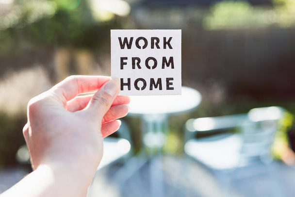 work from home sign being hold in front of sunny out of focus backard with table and chairs, concept of digital nomads working remotely or wfh days during lockdowns or covid-19 isolation - Fotografie, Obrázek
