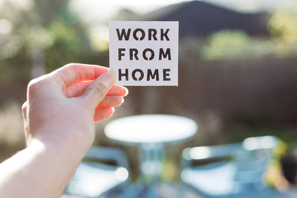 work from home sign being hold in front of sunny out of focus backard with table and chairs, concept of digital nomads working remotely or wfh days during lockdowns or covid-19 isolation - Fotografie, Obrázek