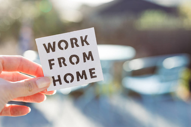 work from home sign being hold in front of sunny out of focus backard with table and chairs, concept of digital nomads working remotely or wfh days during lockdowns or covid-19 isolation - Foto, Imagem