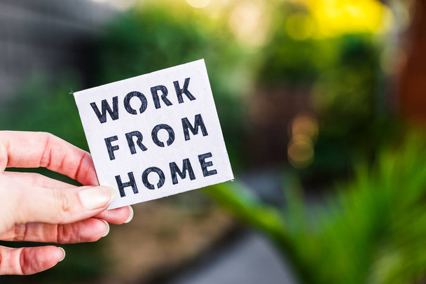 work from home sign being hold in front of out of focus lush green backard, concept of digital nomads working remotely or wfh days during lockdowns or covid-19 isolation - Fotoğraf, Görsel