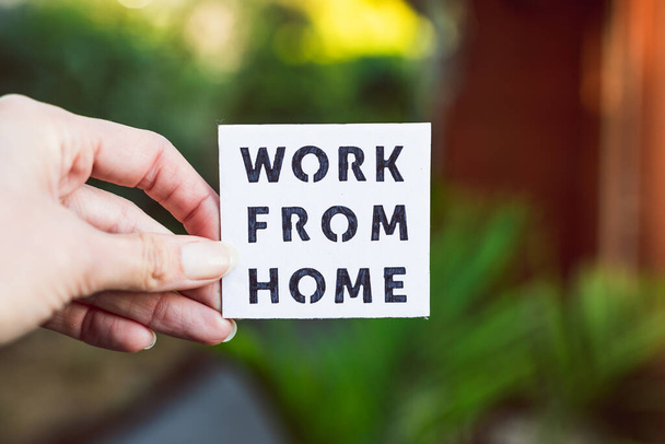 work from home sign being hold in front of out of focus lush green backard and home exterior, concept of digital nomads working remotely or wfh days during lockdowns or covid-19 isolation - Valokuva, kuva