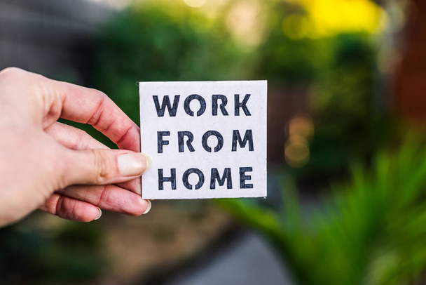 work from home sign being hold in front of out of focus lush green backard and home exterior, concept of digital nomads working remotely or wfh days during lockdowns or covid-19 isolation - Photo, Image