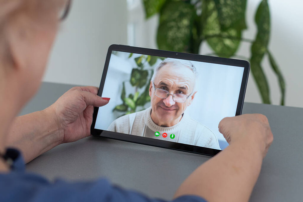 Telegeneeskunde concept, oude vrouw met tablet pc tijdens een online consult met haar arts in haar woonkamer - Foto, afbeelding