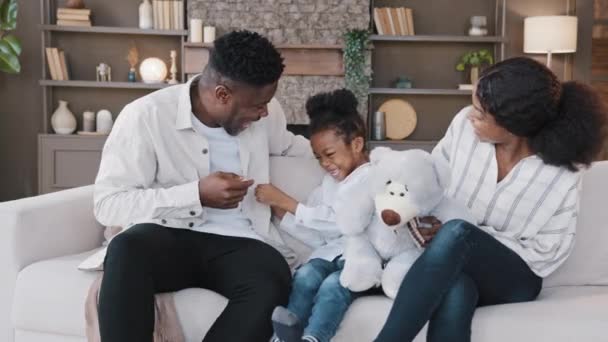 Biracial family sitting on couch in living room spending weekend together. African mother and father with child girl having fun at home. Dad tickling kid daughter on sofa laughing tickle play game - Footage, Video