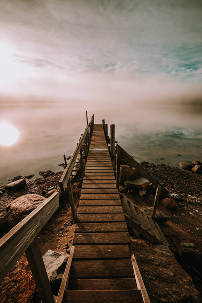 A vertical closeup of a wooden bridge on the lake shore covered in clouds - Фото, зображення