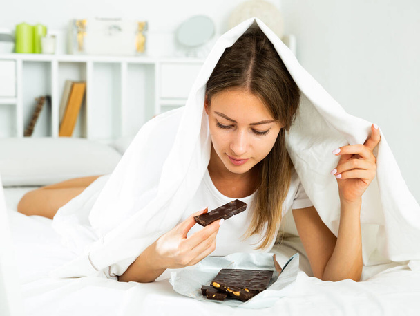 Μια γυναίκα τρώει κρυφά σοκολάτα κάτω από σεντόνια στο κρεβάτι. Υψηλής ποιότητας φωτογραφία - Φωτογραφία, εικόνα