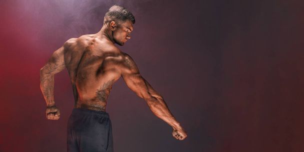 Back view of handsome confident athletic man with bare muscular torso posing on a dark background filled with smoke. Male beauty concept. Fitness, bodybuilding.  - Foto, Imagen