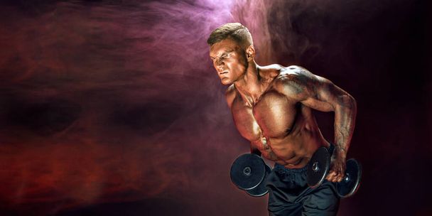Brutal sweaty strong young man athlete with naked upper body standing doing workout with dumbbels and showing strong pumped up biceps over smoky background. Sport men body concept.  - Foto, Imagem