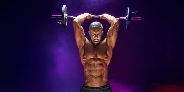Brutal sweaty strong young man athlete with naked upper body standing doing workout with barbell and showing strong pumped up biceps over smoky background. Sport men body concept. - Photo, image
