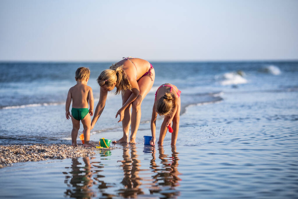 A kind caring beautiful mother plays with her children: eldest skinny daughter and youngest son toddler building beads and sand castles decorating them with shells and pebbles - Foto, Imagem