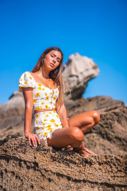 Brunette model in a white dress on the beach enjoying the summer, sitting on some rocks - 写真・画像