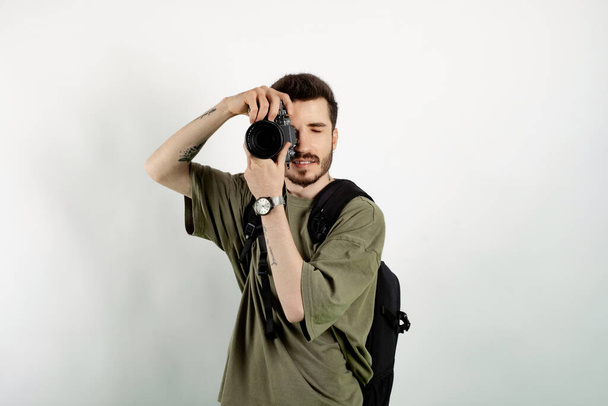 Πορτρέτο του νεαρού όμορφου άνδρα που φοράει χακί t-shirt και ποζάρει απομονωμένος σε λευκό φόντο τραβώντας φωτογραφίες με dslr κάμερα. Φωτογράφος καλύπτει το πρόσωπό του με την κάμερα. - Φωτογραφία, εικόνα