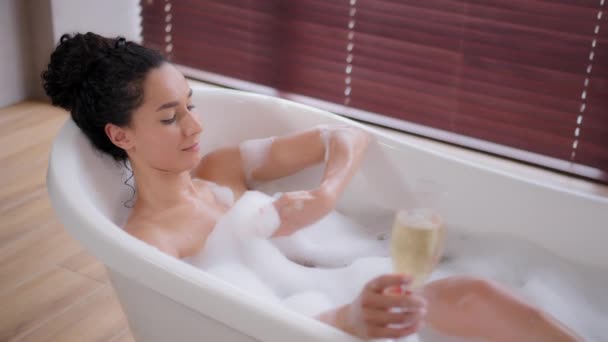 Молодая игривая женщина расслабляется в горячей ванне с пузырьками, держа в руке стеклянное шампанское, наслаждаясь роскошной ванной, романтичная счастливая девушка принимает ванну играет дуя на пены питьевой напиток отдыха - Кадры, видео