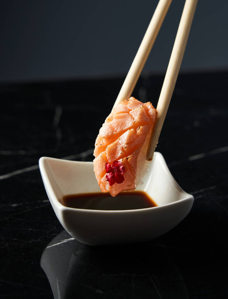 Κοντινό πλάνο Το χέρι κρατάει ξυλάκια μπαμπού με nigiri σολομού, ενώ το χέρι βουτιά nigiri σε σάλτσα σόγιας στο εστιατόριο. Μπροστά. Τρόφιμα, ιαπωνική κουζίνα έννοια. - Φωτογραφία, εικόνα