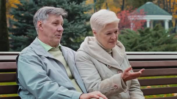 Kaukasische bejaarde familie rusttijd samen in de herfst park zitten op bank getrouwd paar partners senior leeftijd man knuffels volwassen vrouw met grijs kort haar praten zorgeloos buiten roddelen groeten mensen - Video