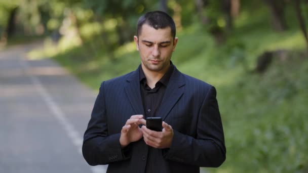 深刻な大人の白人男性ユーザーは、公園で屋外スタンドは、スマートフォンの画面上で携帯電話のスクロールスライドページを保持黒の正式なスーツを身に着けていますオンライン情報テキストメッセージソーシャルビジネスチャット - 映像、動画
