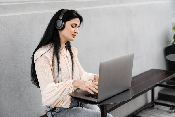 Женщина с витилиго слушать музыку в наушниках и работать на ноутбуке в кафе на открытом воздухе. Руки с пигментацией кожи витилиго. Стиль жизни при сезонных заболеваниях кожи - Фото, изображение