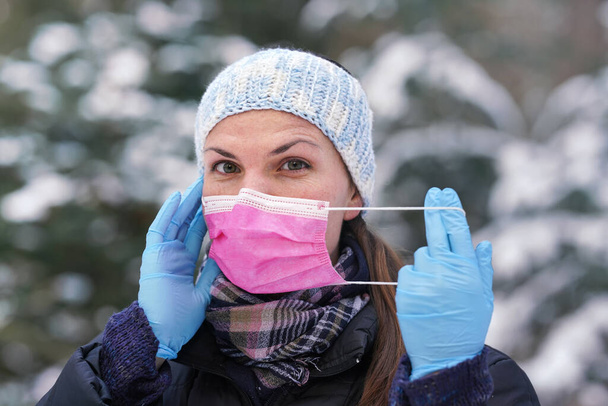 Young woman in warm winter clothes, wearing protection gloves, putting or removing disposable single use pink face virus mouth mask, closeup, blurred snow covered trees background - Φωτογραφία, εικόνα