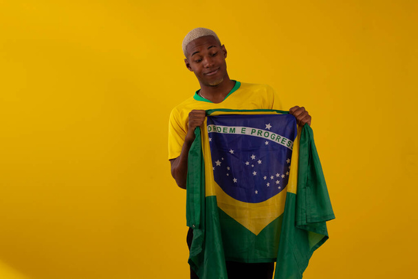 Muž s černou pletí držící brazilskou vlajku a v košili brazilského fotbalového týmu na studiové fotce - Fotografie, Obrázek