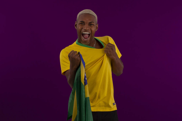 Hombre de piel negra sosteniendo la bandera de Brasil y vistiendo la camisa del equipo de fútbol brasileño en foto de estudio - Foto, Imagen