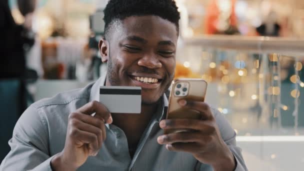 Satisfied african american man makes purchase in online store pays for goods with credit card on mobile phone uses e-banking application happy young male making safe transaction secure payment concept - Footage, Video