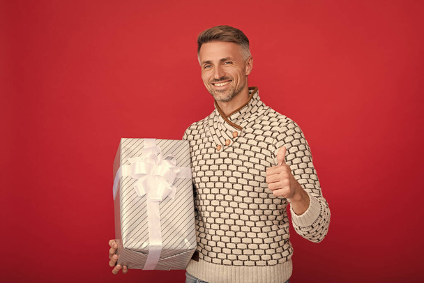 homme heureux en pull tenir boîte cadeau sur fond rouge montrant pouce vers le haut, anniversaire. - Photo, image