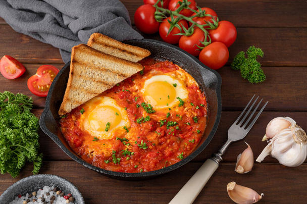 Shakshuka in a frying pan on a dark wooden background. Eggs cooked in tomato sauce with spices and fresh parsley on top. Rustic style. Top view, copy space - Φωτογραφία, εικόνα