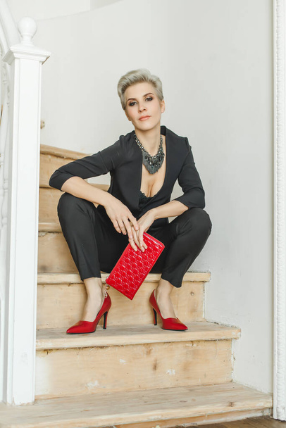 Fashion style portrait of perfect blonde woman in stylish suit with red handbag posing indoor - Foto, Imagen