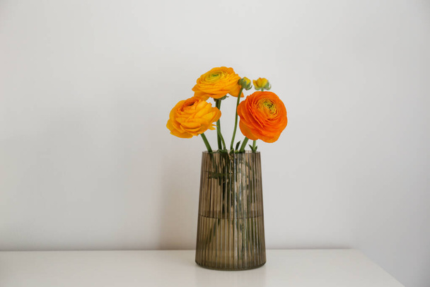 Beautiful tender yellow-orange ranunculus bouquet in a glass vase on table. Visible petal structure. Bright patterns of flower buds. Top view, close up, copy space, white wall background. - Foto, Imagem