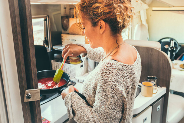 Woman viewed from back preparing and cooking tomato sauce for lunch inside a modern camper van. Off grid and van life lifestyle with traveler people having food preparation. Indoor kitchen activity - Photo, Image