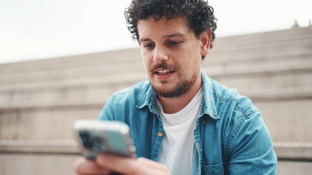smiling young bearded man in denim shirt sitting on high steps and using mobile phone. Man playing on smartphone - Fotó, kép