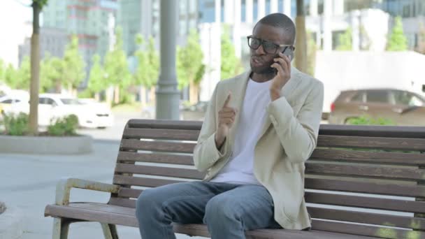 Boos Afro-Amerikaanse man praten over de telefoon tijdens het zitten buiten op de bank - Video