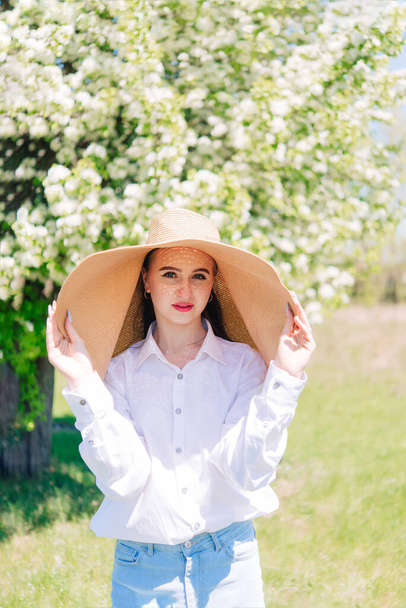 young girl in a straw hat and a white blouse against the backdrop of a flowering tree in spring - Foto, Imagem