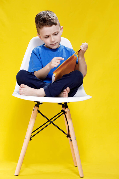 Little cheerful blond green-eyed boy 5-6 years old in a stylish blue T-shirt holding book and reading on yellow wall background, children's studio portrait. - Фото, изображение