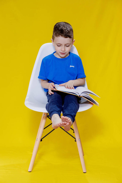 Positive preschool boy in an aspen T-shirt holding an open yellow textbook and looking at it on a yellow background. - Foto, afbeelding