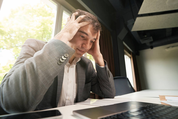 Male office employee suffering from too much workload and holding his head while staring at paper sheet - Foto, Imagem