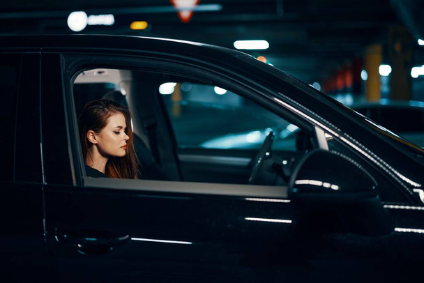 foto horizontal desde un lado, por la noche, de una mujer sentada en un coche negro y mirando pensativamente hacia adelante. Foto de alta calidad - Foto, Imagen