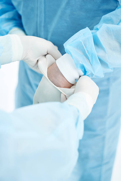 Hemşirenin ameliyat önlüğü giymiş tanınmayan bir doktorun eline lateks eldiven takmasına yakın çekim. - Fotoğraf, Görsel