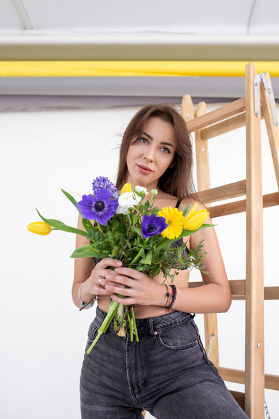 Bouquet of spring flowers. Beautiful young woman in dark gray jeans with dreamy look holds flowers in her hands, standing in studio on white background. Romantic mood. Delivery of bouquets.  - Фото, изображение
