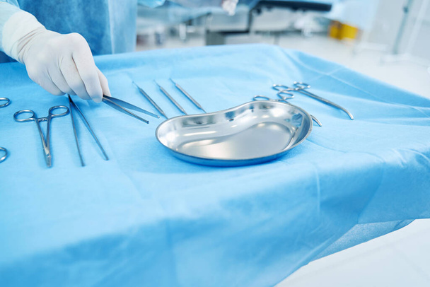 Close up of a table with surgical instruments. Hand in latex glove picking up forceps - Photo, Image